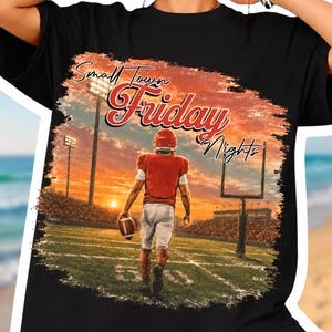 Könnte beinhalten: Ein schwarzes T-Shirt mit einem Grafikdruck eines Footballspielers in rotem Trikot und weißer Hose auf einem Spielfeld bei Sonnenuntergang. Der Text "Small Town Friday Nights" ist in einer stilisierten rot-schwarzen Schrift abgebildet.