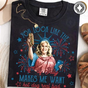 May include: Black t-shirt with a graphic of a woman dressed as the Statue of Liberty holding a hot dog. The text reads "You look like the 4th of July makes me want hot dog real bad." A gold necklace is draped on the shirt.