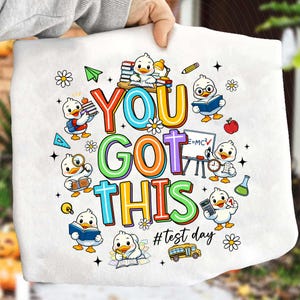 Peut inclure: Tissu blanc avec un graphisme coloré indiquant "YOU GOT THIS" avec des canetons de dessins animés tenant des livres, des crayons et une calculatrice. La phrase "#test day" est également incluse. Le motif est entouré de marguerites, d'étoiles et d'un bus scolaire.