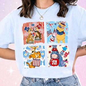 May include: Pale blue t-shirt with four square graphics of Disney characters celebrating the 4th of July. Chip and Dale, Winnie the Pooh, Piglet, Tigger, and Eeyore are shown with flags and fireworks.