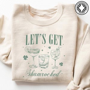 Könnte beinhalten: Cremefarbenes Sweatshirt mit dem grünen Text "LET'S GET Shamrocked". Das Design zeigt Illustrationen von drei Cocktails, Kleeblättern und Sternen. Ein lustiges, festliches Oberteil für St. Patrick's Day oder jede Feier.