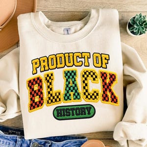 Pode incluir: Camisola creme com o texto "PRODUCT OF BLACK HISTORY" em letras xadrez amarelas, pretas, verdes e vermelhas. A camisola está dobrada e o texto está centrado na frente.