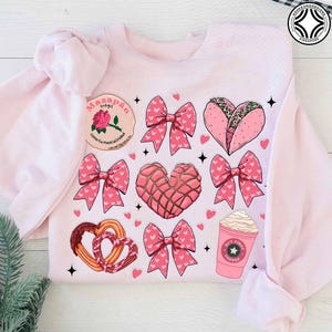 May include: Light pink sweatshirt with a pattern of pink bows, hearts, and food illustrations. The illustrations include a heart-shaped taco, a concha, churros, and a coffee cup with the text "BUT FIRST COFFEE". A patch with text is also visible.
