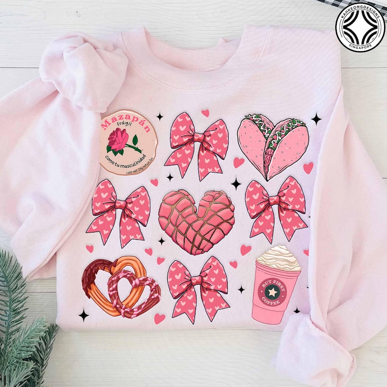 May include: Light pink sweatshirt with a pattern of pink bows, hearts, and food illustrations. The illustrations include a heart-shaped taco, a concha, churros, and a coffee cup with the text "BUT FIRST COFFEE". A patch with text is also visible.