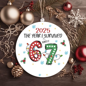 Könnte beinhalten: Ein runder Weihnachtsschmuck mit dem Text "2025 The Year I Survived Barely". Die Zahlen "67" sind mit Lichterketten verziert. Der Schmuck ist weiß und hat eine Schnur zum Aufhängen.