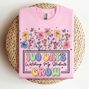 Pode incluir: Camisola rosa claro com um design floral e o texto "100 Days Watching My Students GROW" em uma fonte retr&ocirc; colorida. O design inclui v&aacute;rias flores e borboletas coloridas.