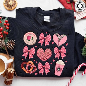May include: Black sweatshirt with a variety of pink graphics, including bows, hearts, a taco, a coffee cup, and a pretzel. The sweatshirt also features a circular patch with text and a rose. The design is festive and playful.