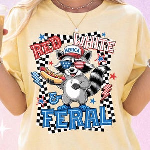 Può includere: T-shirt gialla con un procione dei cartoni animati con occhiali da sole e un berretto da baseball con la scritta "Merica". Il procione tiene in mano un hot dog e il testo "Red White & Feral" è visualizzato in una cornice a scacchi.