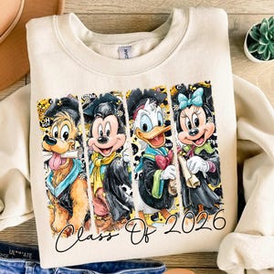 Puede incluir: Sudadera color crema con cuatro personajes de dibujos animados: Pluto, Mickey Mouse, Donald Duck y Minnie Mouse, cada uno con atuendo de graduación. El texto "Class Of 2026" está escrito en una fuente cursiva en la parte inferior.