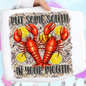 May include: White tote bag featuring a graphic of two red crawfish, yellow lemon slices, and the text "PUT SOME SOUTH IN YOUR MOUTH." The design has a distressed, vintage look with a camouflage-style background.