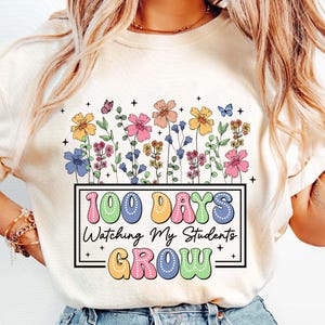 Pode incluir: Camiseta creme com um design floral colorido e borboletas. A camiseta tem o texto "100 DAYS Watching My Students GROW" em uma moldura retangular. O texto e as flores s&atilde;o de v&aacute;rias cores.