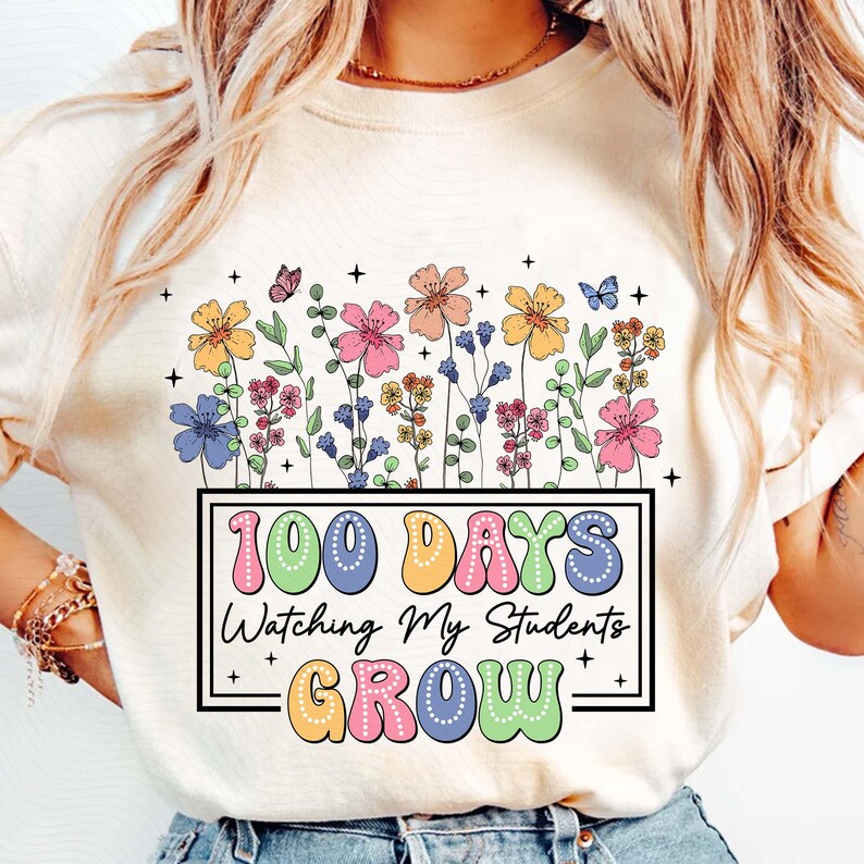 Pode incluir: Camiseta creme com um design floral colorido e borboletas. A camiseta tem o texto "100 DAYS Watching My Students GROW" em uma moldura retangular. O texto e as flores s&atilde;o de v&aacute;rias cores.