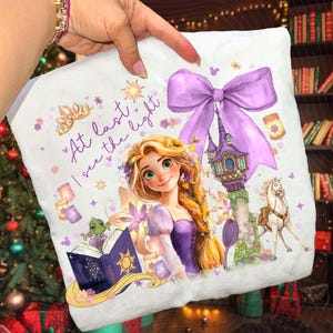 May include: White decorative pillow featuring a princess illustration with the text "At last I see the light." The design includes a castle, horse, and a large purple bow. The pillow has a soft, plush texture.