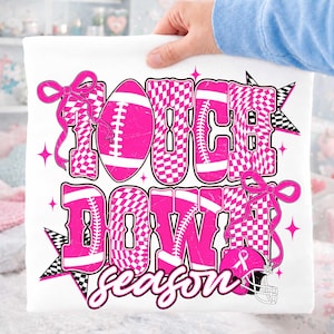 May include: A white t-shirt featuring a vibrant pink and white checkered graphic with the text TOUCH DOWN season. The design includes footballs, a football helmet with a breast cancer awareness ribbon, and pink bows, accented by black and white stars.