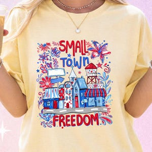 Könnte beinhalten: Hellgelbes T-Shirt mit einem farbenfrohen Grafikdesign einer Kleinstadt. Das Design umfasst Gebäude, Blumen und die Worte "SMALL TOWN FREEDOM" in Rot und Blau. Das T-Shirt besteht aus einem weichen Material.
