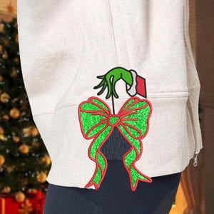 May include: A beige hoodie with a green and red Christmas bow design. The design features a green hand holding a red and white gift tag above the bow. The bow is green with red trim.