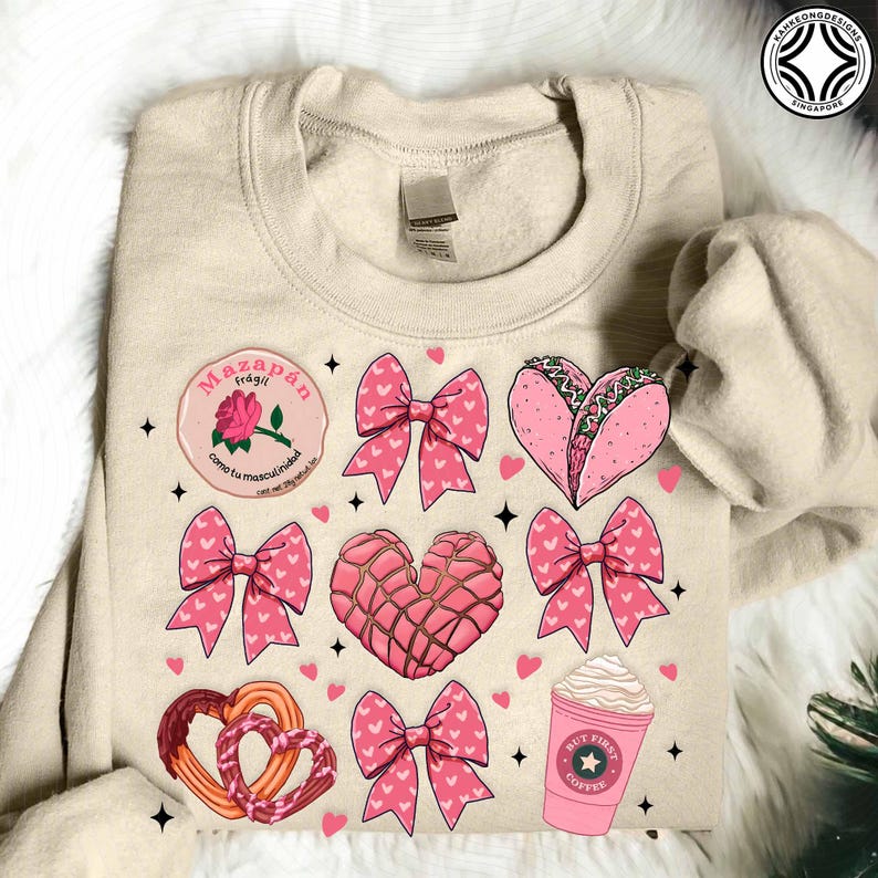May include: Beige sweatshirt with a variety of pink graphics, including bows, hearts, a taco, a coffee cup, and a rose. The sweatshirt has the text "Mazap&aacute;n fr&aacute;gil" and "como tu masculinidad".