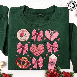 May include: Dark green sweatshirt featuring a variety of pink and red graphics. Designs include bows, hearts, a taco, a coffee cup, and a pretzel. A circular patch reads "Mazap&aacute;n fr&aacute;gil como tu masculinidad".