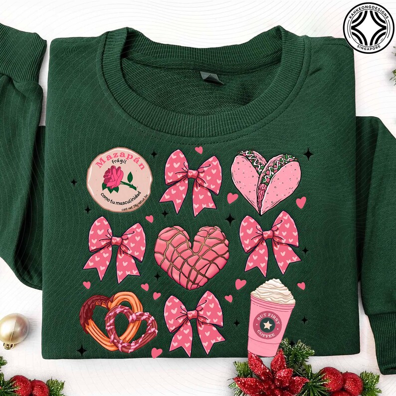 May include: Dark green sweatshirt featuring a variety of pink and red graphics. Designs include bows, hearts, a taco, a coffee cup, and a pretzel. A circular patch reads "Mazap&aacute;n fr&aacute;gil como tu masculinidad".