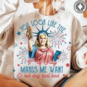 May include: White sweatshirt with a graphic featuring a woman dressed as the Statue of Liberty holding a hot dog. Text includes "You look like the 4th of July makes me want hot dog real bad" in red and blue.
