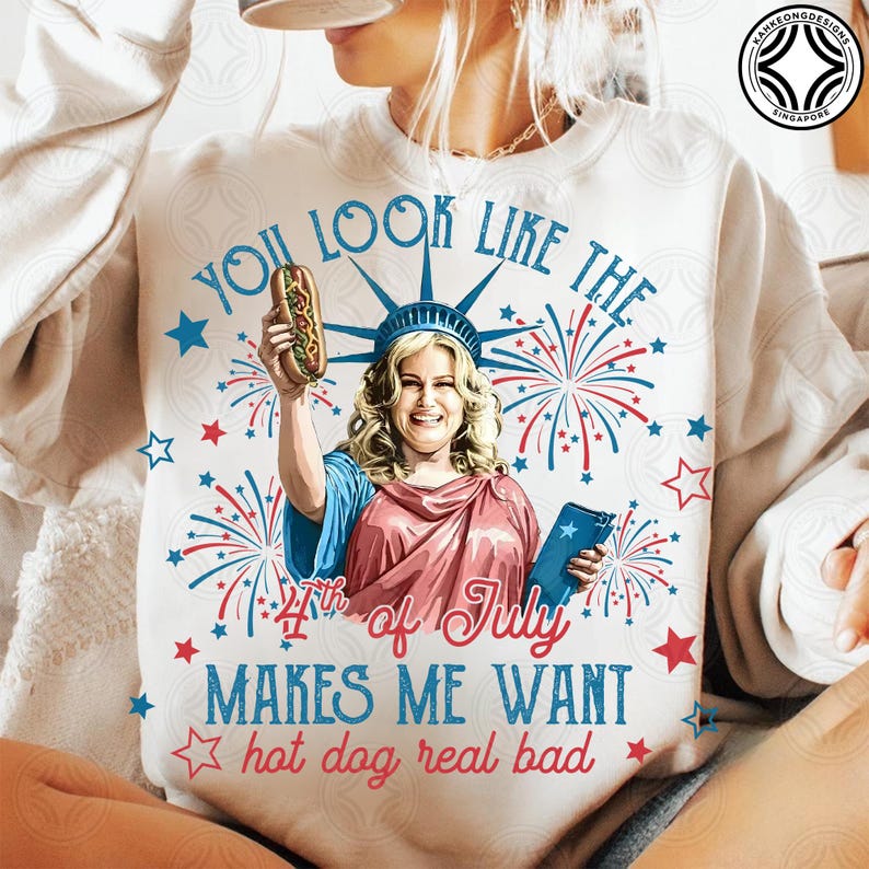 May include: White sweatshirt with a graphic featuring a woman dressed as the Statue of Liberty holding a hot dog. Text includes "You look like the 4th of July makes me want hot dog real bad" in red and blue.
