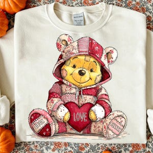 Könnte beinhalten: Cremefarbenes Sweatshirt mit einer Illustration eines Teddybären in einem Patchwork-Kapuzenpullover, der ein rotes Herz mit "LOVE" hält. Auf den Füßen des Bären steht "HUGS" und "KISSES".