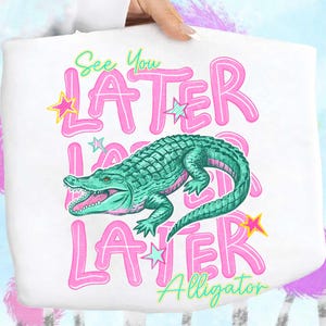 May include: White tote bag featuring a teal alligator illustration with the words "See You LATER Alligator" in pink and green. The design includes star accents, creating a playful and vibrant aesthetic.