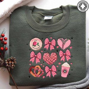 May include: An olive green sweatshirt featuring a collection of pink and white graphics. The graphics include bows, hearts, a taco, a coffee cup, and a round emblem with text. The sweatshirt is a casual, comfortable garment.