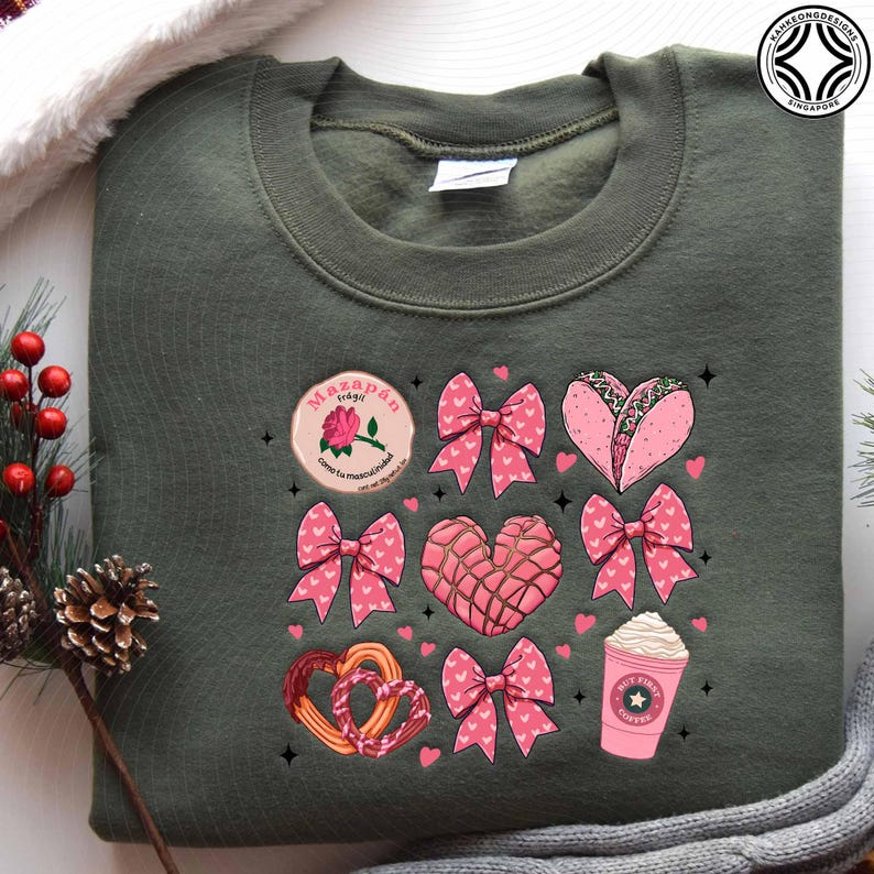 May include: An olive green sweatshirt featuring a collection of pink and white graphics. The graphics include bows, hearts, a taco, a coffee cup, and a round emblem with text. The sweatshirt is a casual, comfortable garment.