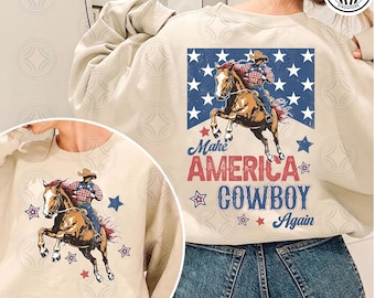 May include: A beige sweatshirt featuring a graphic of a cowboy riding a horse, with the text "Make America Cowboy Again" in red and blue. The design includes stars and a vintage aesthetic, suitable for Western-themed apparel.