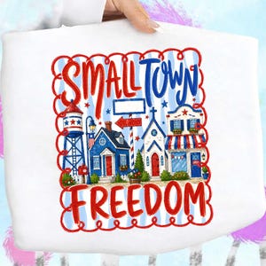 Könnte beinhalten: Weiße Tragetasche mit einer farbenfrohen Illustration einer Kleinstadt. Das Design umfasst einen Wasserturm, Häuser und die Worte "SMALL TOWN FREEDOM" in Rot und Blau. Der Hintergrund hat blau-weiße Streifen.