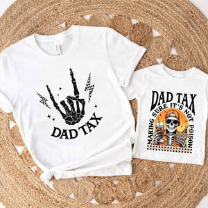 May include: Two white t-shirts with black graphics. One shirt features a skeleton hand making a rock and roll gesture with the text "DAD TAX". The other shirt has a skeleton with a drink and fries, with the text "DAD TAX, MAKING SURE IT'S NOT POISON".