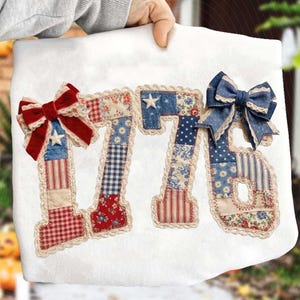 May include: A white fabric item featuring the number "1776" appliqued with patriotic fabric patterns. Each number is trimmed with lace and adorned with bows and star accents in red, white, and blue.
