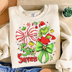 May include: Cream-colored sweatshirt featuring a festive Grinch design. The Grinch is wearing a Santa hat and red suit, with a green bow and the text "Six Seven". The design includes bows, bells, and snowflakes.