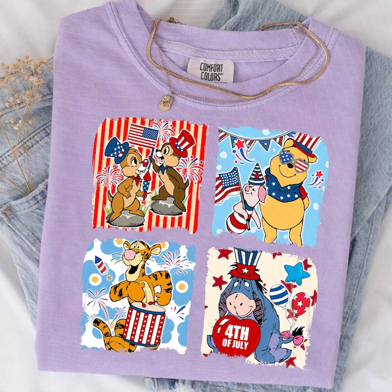 May include: A lilac Comfort Colors t-shirt with four square graphics of patriotic Disney characters. Chip and Dale hold flags, Winnie the Pooh wears sunglasses, Tigger plays a drum, and Eeyore holds a heart that says "4th of July".