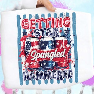 May include: White tote bag with a patriotic design. The design features three beverage cans with American flag motifs, the words "GETTING STAR SPANGLED HAMMERED" in red and blue, and star and firework accents. The bag is held by a person.