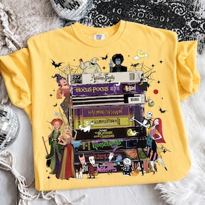 May include: A yellow t-shirt featuring a graphic print of a stack of VHS tapes labeled The Addams Family, Hocus Pocus, Halloweentown, Halloweentown II, The Nightmare Before Christmas, and Goosebumps, surrounded by various illustrated movie characters.