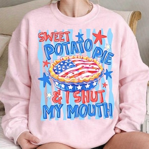 May include: Light pink sweatshirt with a graphic of a sweet potato pie decorated with an American flag design. The text reads "Sweet Potato Pie & I Shut My Mouth" in red and blue.