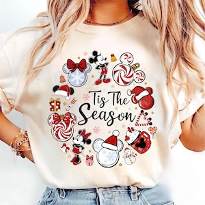 May include: Cream-colored t-shirt featuring a festive Christmas wreath design with Mickey and Minnie Mouse characters. The wreath includes candy canes, snowflakes, and the text "Tis The Season" in black script. The design incorporates red, white, and black colors.