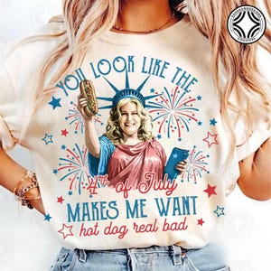 May include: Cream-colored t-shirt with a graphic featuring a woman dressed as the Statue of Liberty holding a hot dog. Text includes "You look like the 4th of July makes me want hot dog real bad" with star and firework accents.