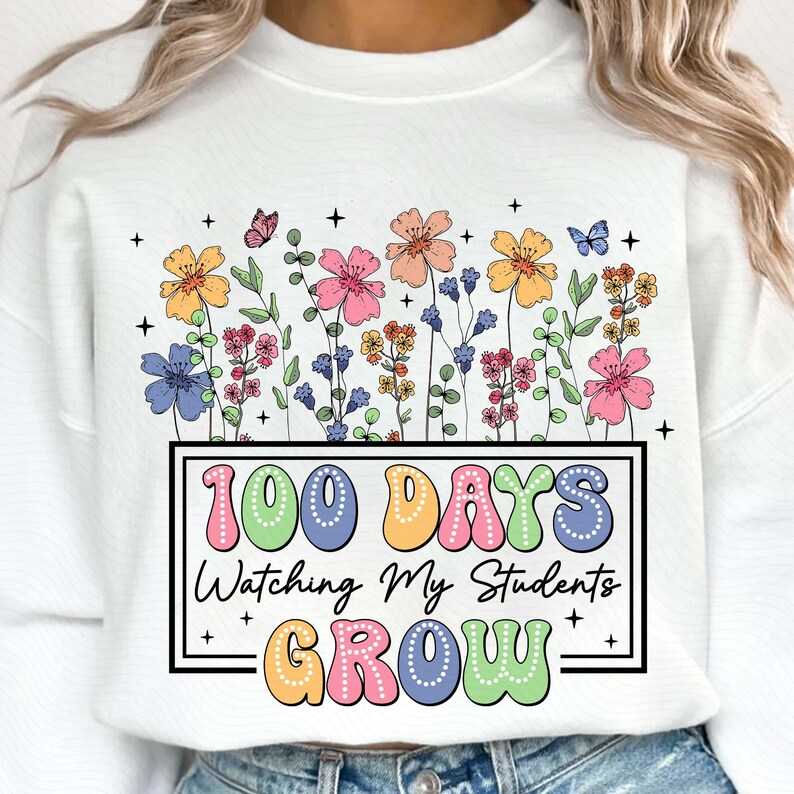 Pode incluir: Camisola branca com um design floral colorido e o texto "100 DAYS Watching My Students GROW" em uma fonte divertida. O design apresenta flores em tons de amarelo, rosa, azul e laranja, com borboletas e pequenas cruzes.