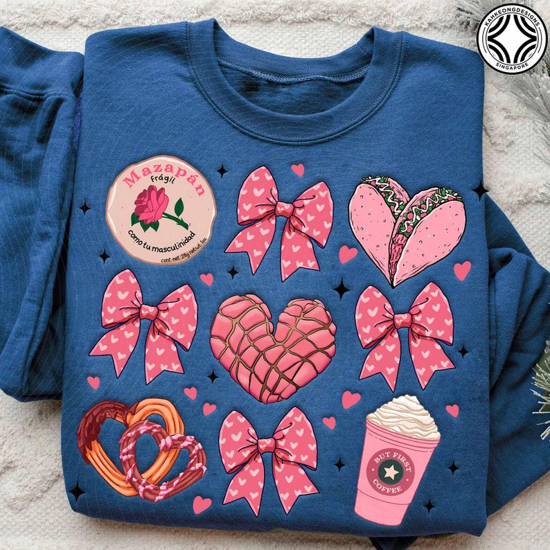 May include: A blue sweatshirt featuring various pink graphics, including bows, hearts, a taco, a coffee cup, and a pastry. The sweatshirt also has text in a circle that says "Mazap&aacute;n Fr&aacute;gil" and "como tu masculinidad".