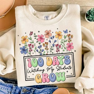 Pode incluir: Camisola creme com um design floral e o texto "100 Days Watching My Students Grow". O gr&aacute;fico apresenta flores coloridas, borboletas e estrelas. A camisola &eacute; feita de um material macio.
