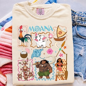 May include: A cream-colored t-shirt featuring a graphic print of Moana characters including Hei Hei, Pua, Maui, and Moana, along with a sailboat, coconut drink, stingray, and Kakamora. The text Moana is printed in blue at the top of the design.