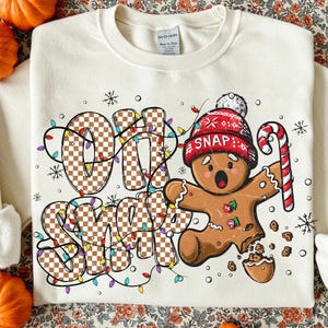 May include: Cream-coloured sweatshirt with a festive design. The graphic features the words "OH SNAP" in a checkered pattern, a gingerbread man wearing a red hat, and a candy cane. The design is accented with string lights and snowflakes.