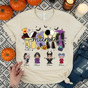 Peut inclure: T-shirt beige avec le mot "Mama" dans un design sur le th&egrave;me d'Halloween. Le motif comprend des chauves-souris, des &eacute;toiles et des images de dessins anim&eacute;s de m&eacute;chants Disney. Des citrouilles et des bougies entourent le t-shirt.