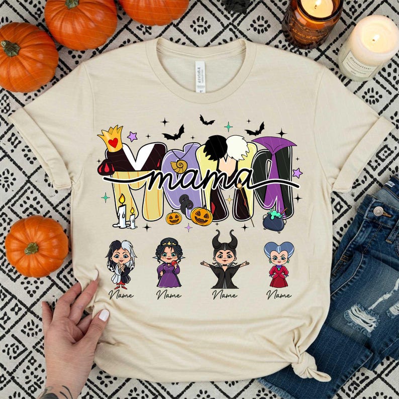 Peut inclure: T-shirt beige avec le mot "Mama" dans un design sur le th&egrave;me d'Halloween. Le motif comprend des chauves-souris, des &eacute;toiles et des images de dessins anim&eacute;s de m&eacute;chants Disney. Des citrouilles et des bougies entourent le t-shirt.
