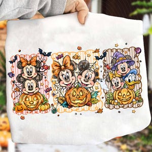 May include: A white t-shirt featuring three rectangular panels with Disney characters in Halloween themes. The panels show Mickey and Minnie Mouse with jack-o'-lanterns, spiderwebs, bats, and candy, plus Donald Duck in a witch hat.