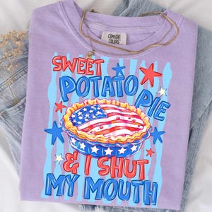 May include: A lilac-colored t-shirt with the words "Sweet Potato Pie & I Shut My Mouth" in a patriotic design. The graphic includes a pie with an American flag design, stars, and blue stripes. A gold necklace is draped over the shirt.