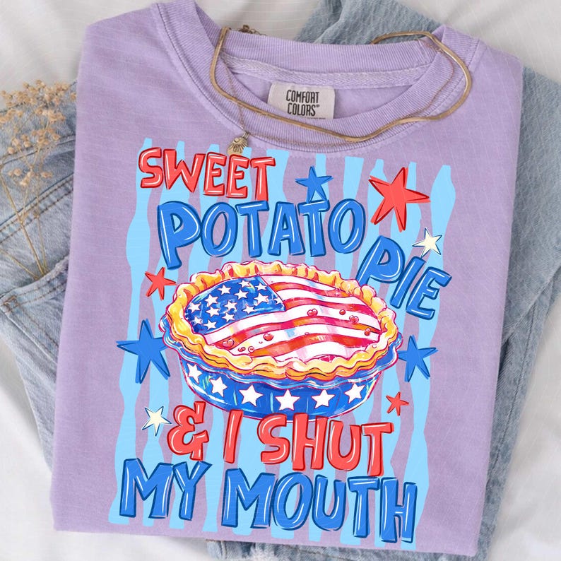 May include: A lilac-colored t-shirt with the words "Sweet Potato Pie & I Shut My Mouth" in a patriotic design. The graphic includes a pie with an American flag design, stars, and blue stripes. A gold necklace is draped over the shirt.